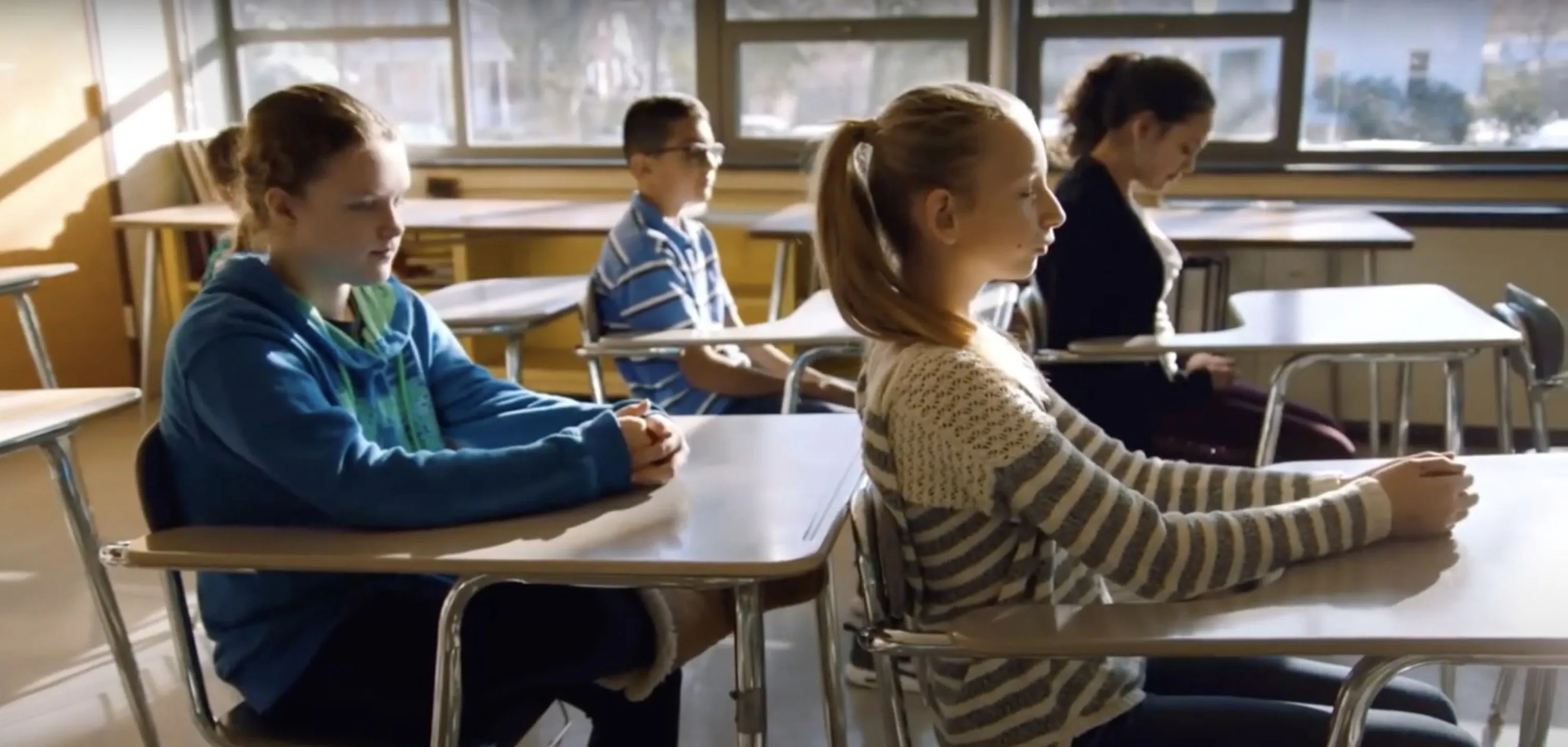 Students sit upright at their desks with eyes closed during a guided mindfulness practice in a sunlit classroom